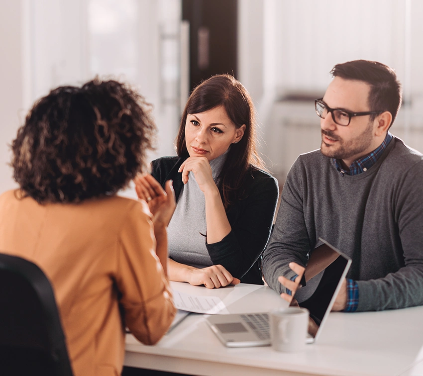 Three people sit at a table having a discussion; two face the camera, listening intently, while the third, back to the camera, speaks. Laptops, papers, and a mug are on the table in a bright office setting.