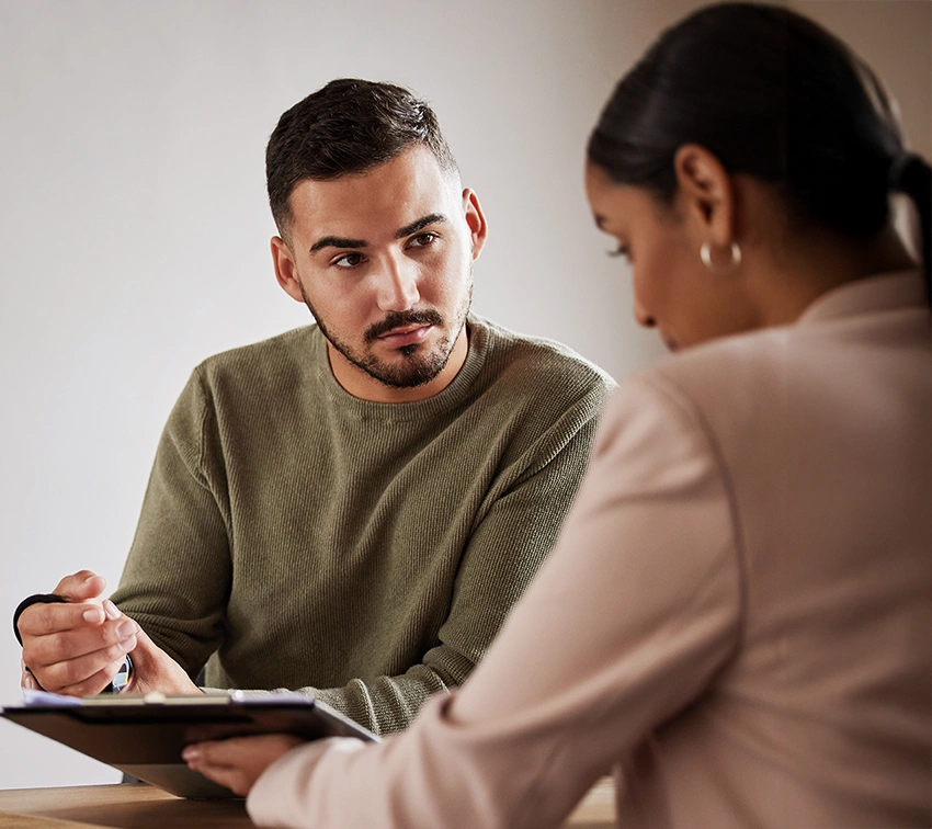 A man in a green sweater attentively listens to a woman in a beige blazer, who holds a clipboard and pen. They are seated at a table, engaged in a serious conversation or discussion.