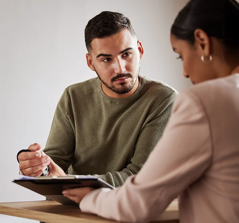 A man in a green sweater attentively listens to a woman in a beige blazer, who holds a clipboard and pen. They are seated at a table, engaged in a serious conversation or discussion.