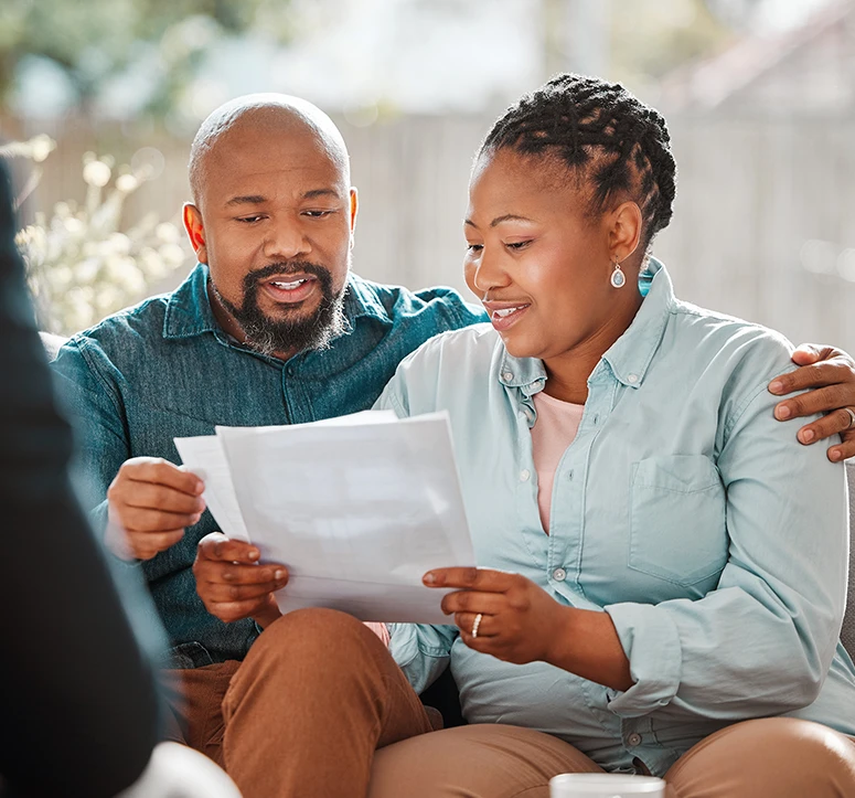 A smiling couple sits together, reviewing paperwork, while another person, partially visible in the foreground, appears to be assisting them. The atmosphere is bright and positive.