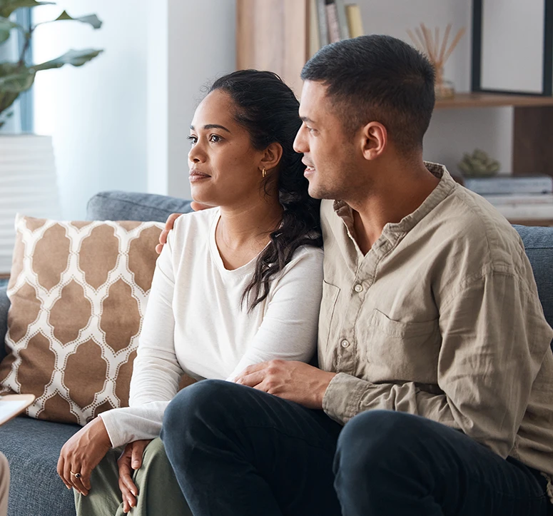 A couple sits closely together on a couch, attentively listening to a lawyer in a bright room with a plant and decorative cushions in the background.