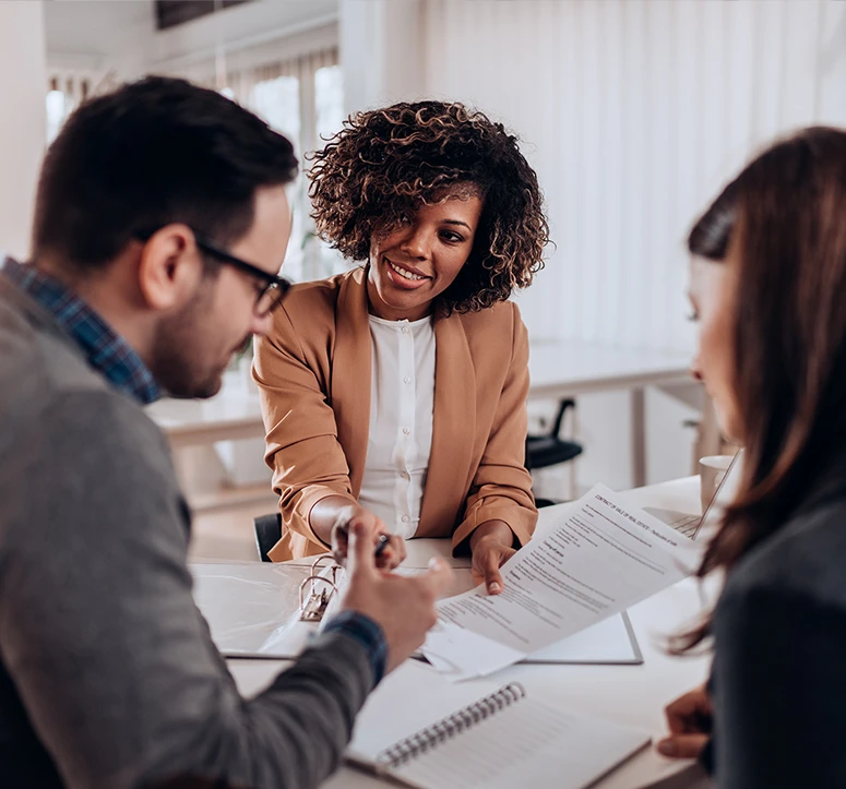 Three people sit at a table in an office. A smiling woman in a tan blazer holds a document and gestures toward it, while the other two people listen and review the paperwork.