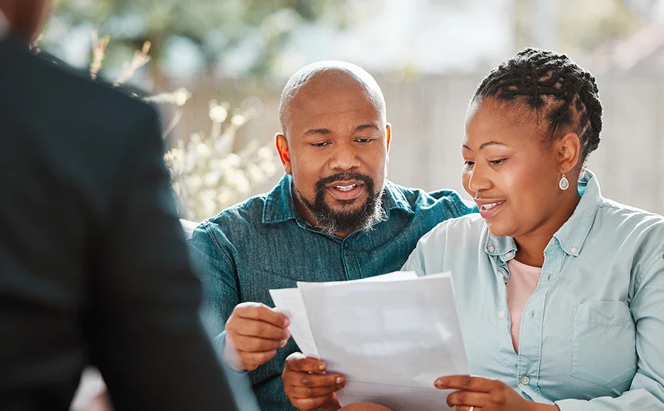 A smiling couple sits together, reviewing paperwork, while another person, partially visible in the foreground, appears to be assisting them. The atmosphere is bright and positive.