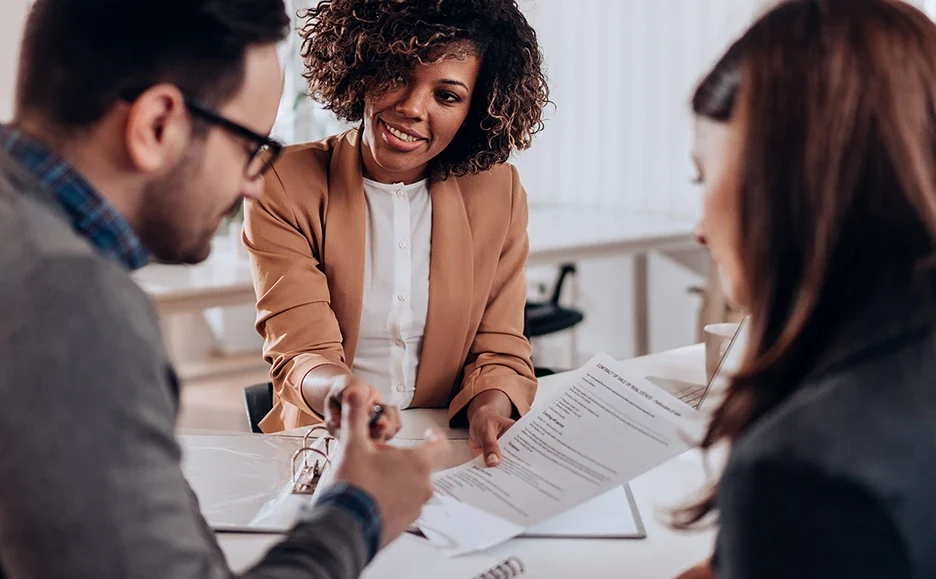 Three people sit at a table in an office. A smiling woman in a tan blazer holds a document and gestures toward it, while the other two people listen and review the paperwork.