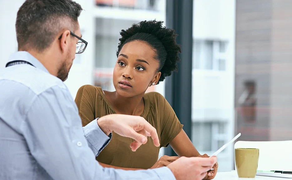 A woman attentively listens to a man holding papers while they have a discussion at a desk with a tablet and a coffee mug in an office setting.