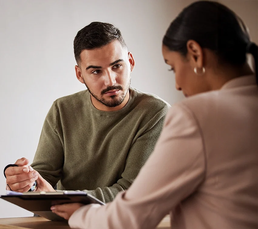A man in a green sweater attentively listens to a woman in a beige blazer, who holds a clipboard and pen. They are seated at a table, engaged in a serious conversation or discussion.
