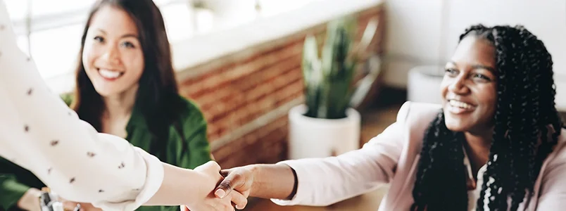 Two women sitting at a table, smiling, as one reaches out to shake hands with a person standing in front of them. There is a potted plant and a large window in the background.