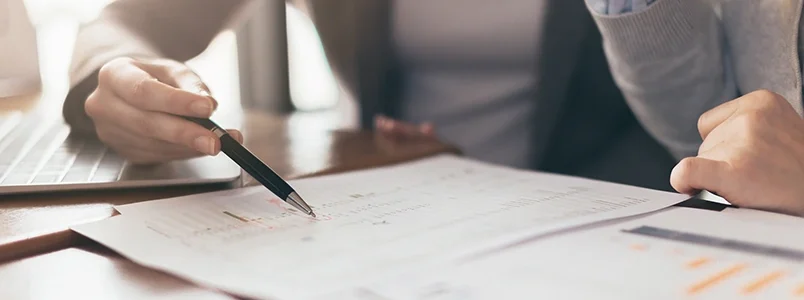 Two people sit at a desk reviewing documents with charts and graphs; one person points at the papers with a pen while the other looks on, and a laptop is partially visible in the background.