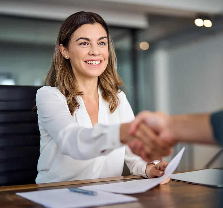 A smiling woman in a white blouse sits at a desk holding papers and shaking hands with someone out of frame, suggesting a successful meeting or job interview.