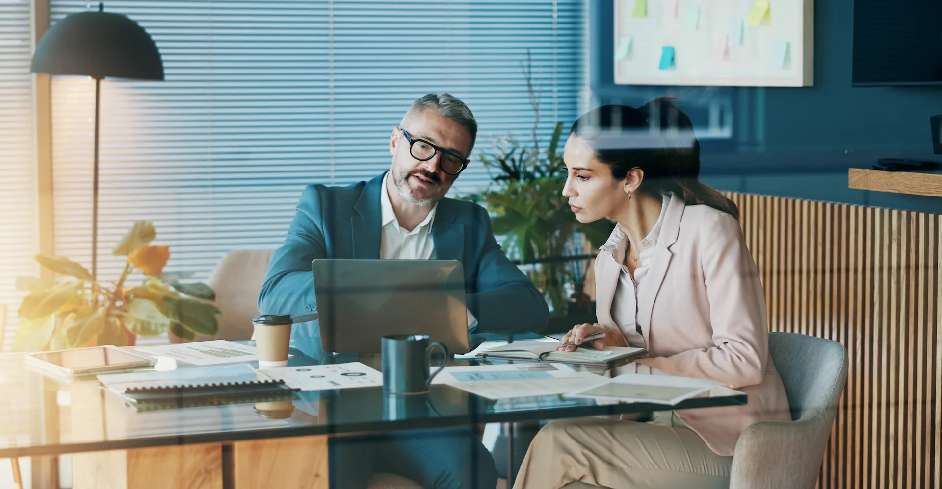 Two business professionals sit at a table in a modern office, reviewing documents and working on a laptop. Both appear focused and engaged in discussion, with coffee cups and papers on the table.