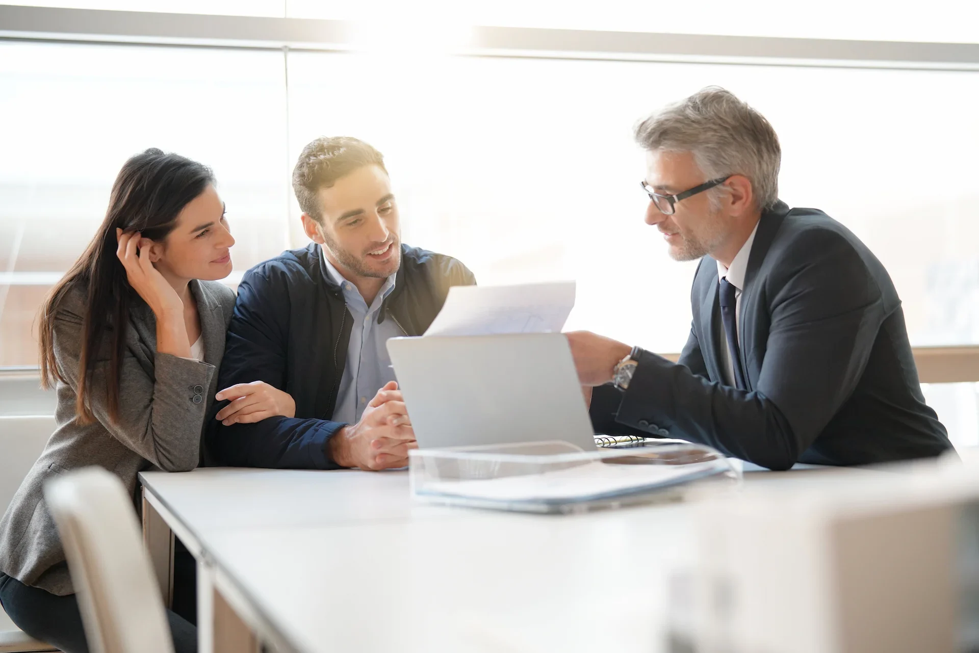 A professional man in a suit shows documents to a smiling couple sitting across from him at a desk in a bright office setting, suggesting a business meeting or consultation.