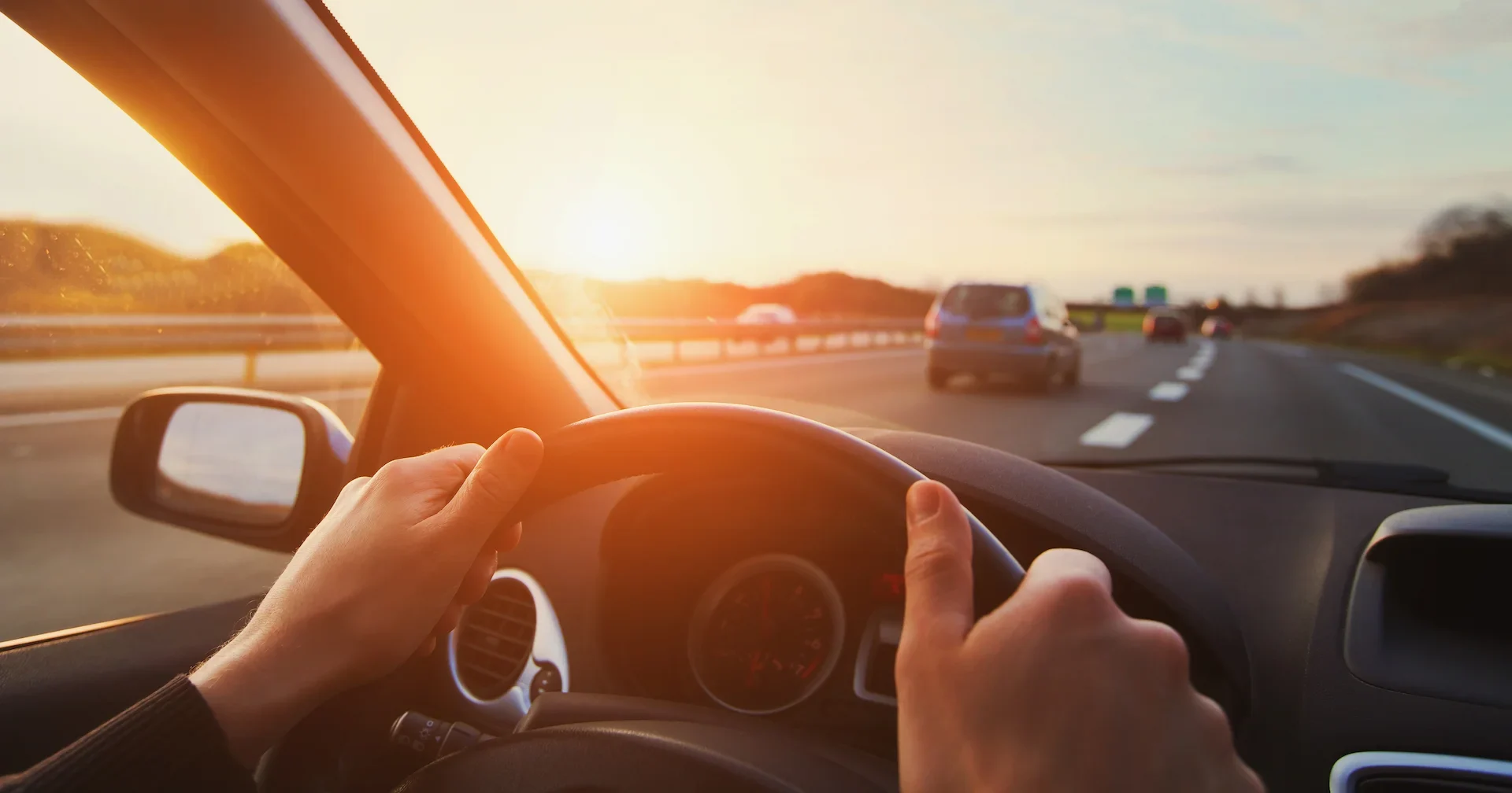 A person’s hands on a steering wheel driving a car on a highway at sunset, with other cars visible ahead and the sun low on the horizon.