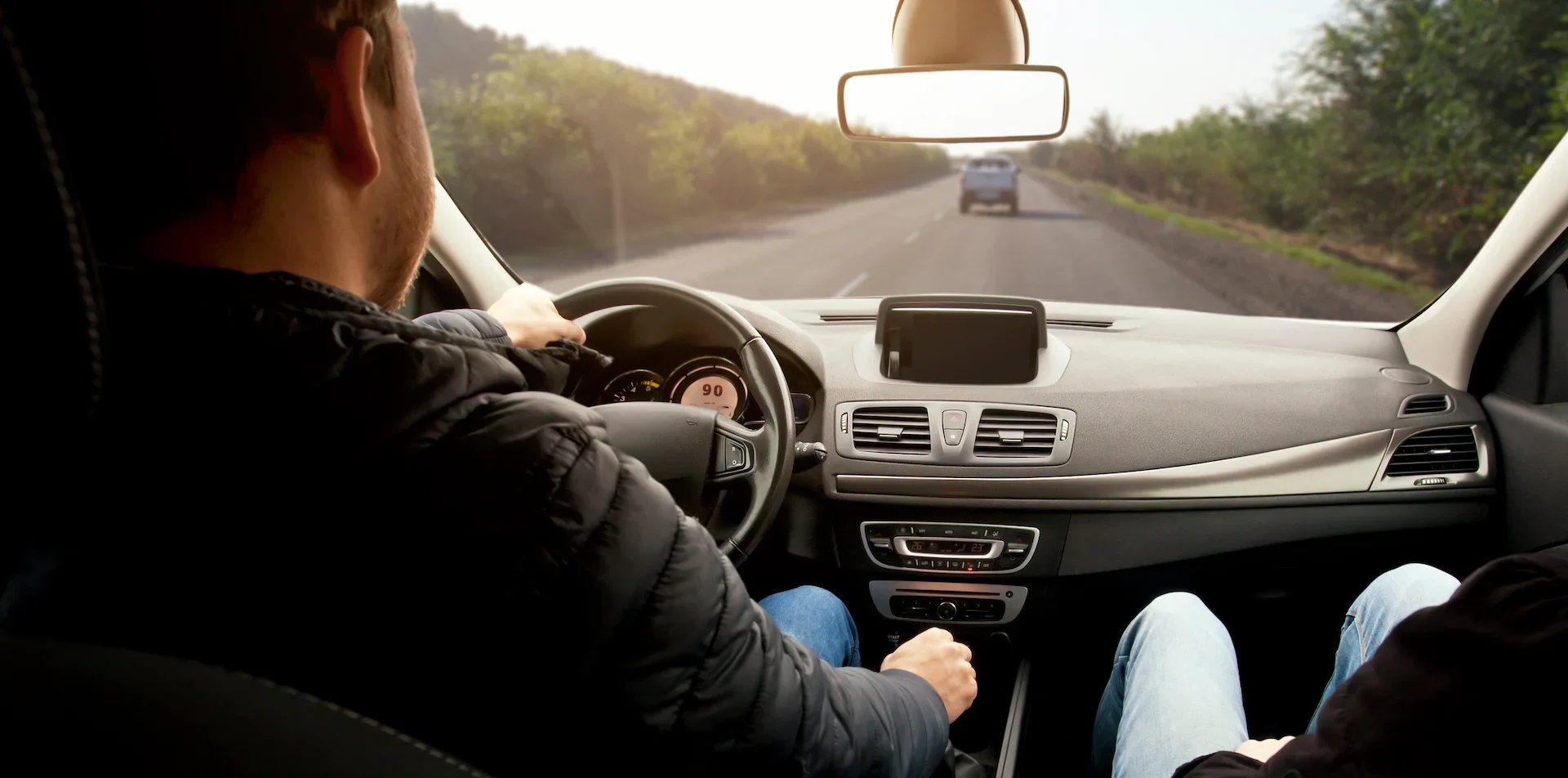 A person is driving a car on a two-lane road with another passenger in the front seat; the dashboard and road ahead are visible through the windshield.