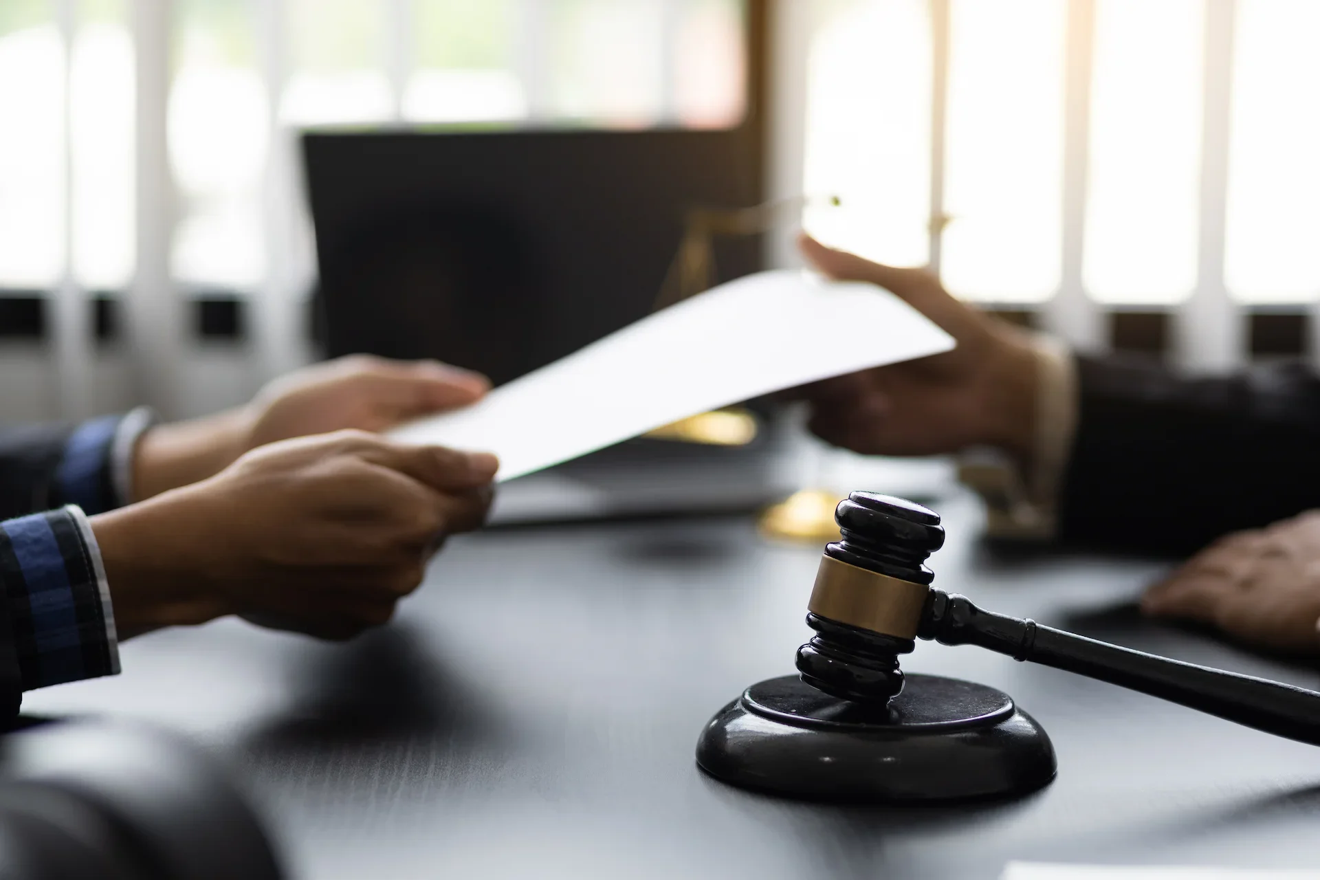 Two people sitting at a desk, exchanging a document; a judges gavel is prominently placed in the foreground, suggesting a legal or courtroom setting.