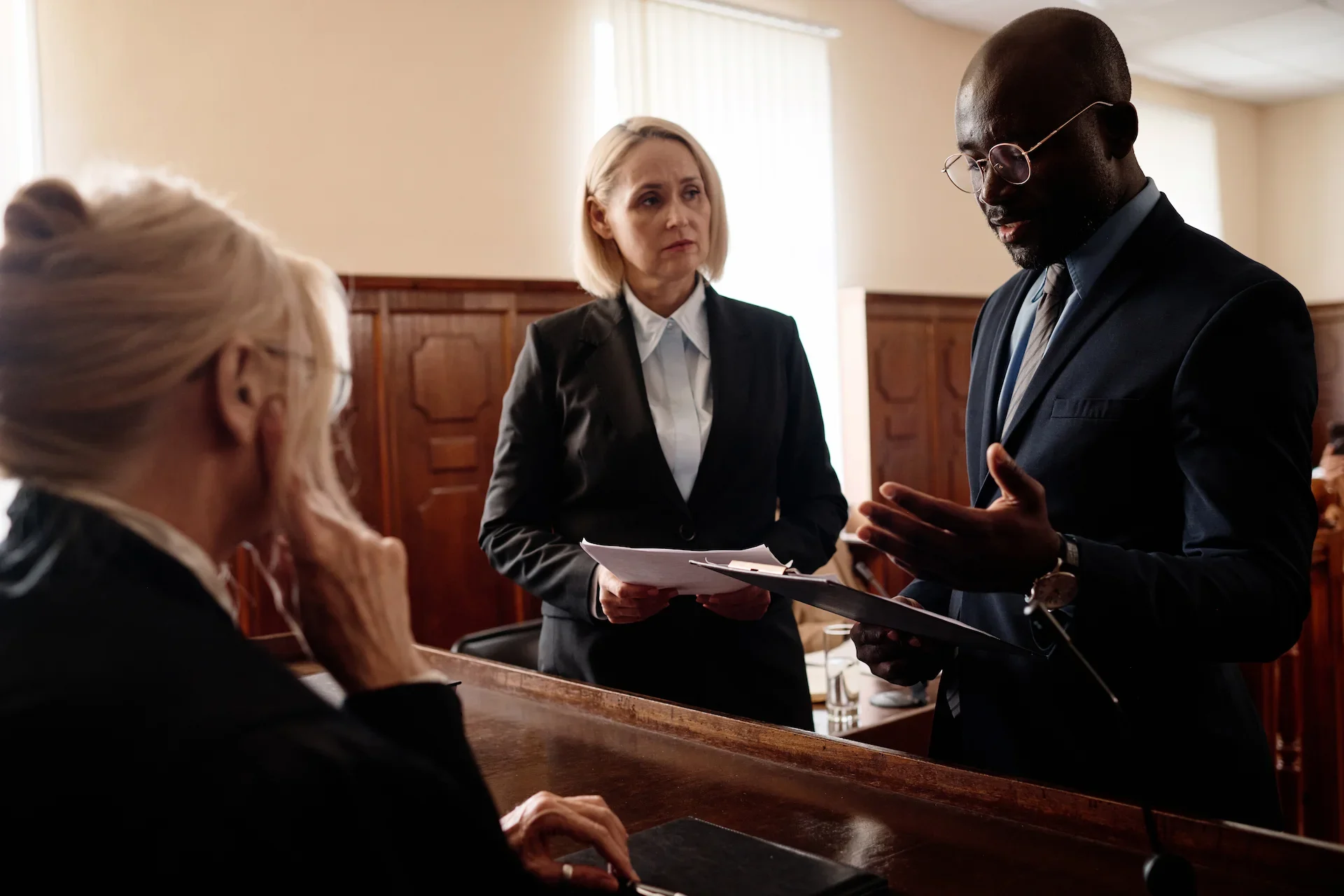 A man in a suit speaks while holding papers in a courtroom, with a woman in a suit standing beside him and another woman, possibly a judge, seated across from them.