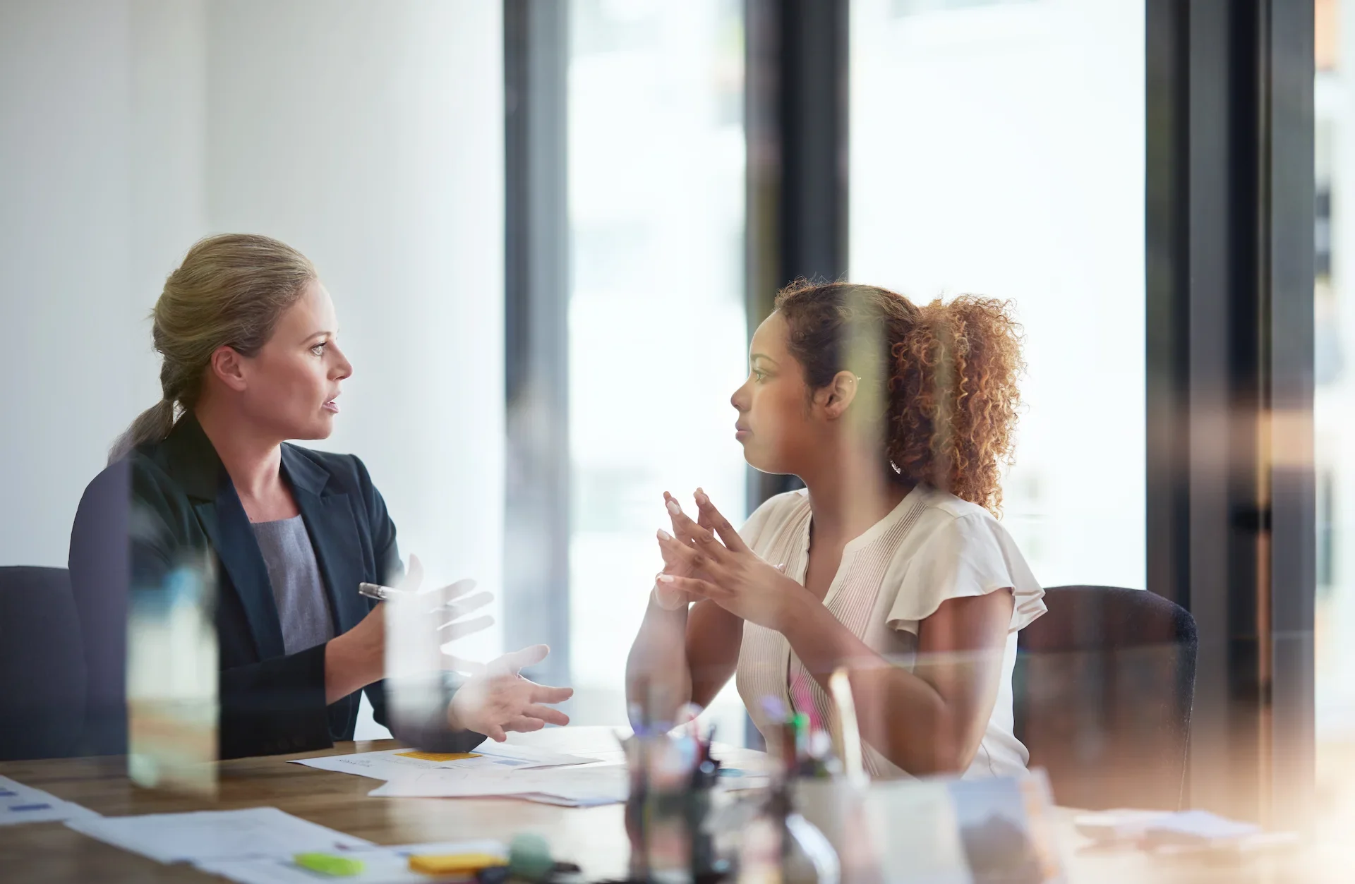 Two women sit at a table in an office, having a serious conversation. Papers and pens are on the table, and sunlight streams through large windows behind them. Both women appear focused and engaged in discussion.